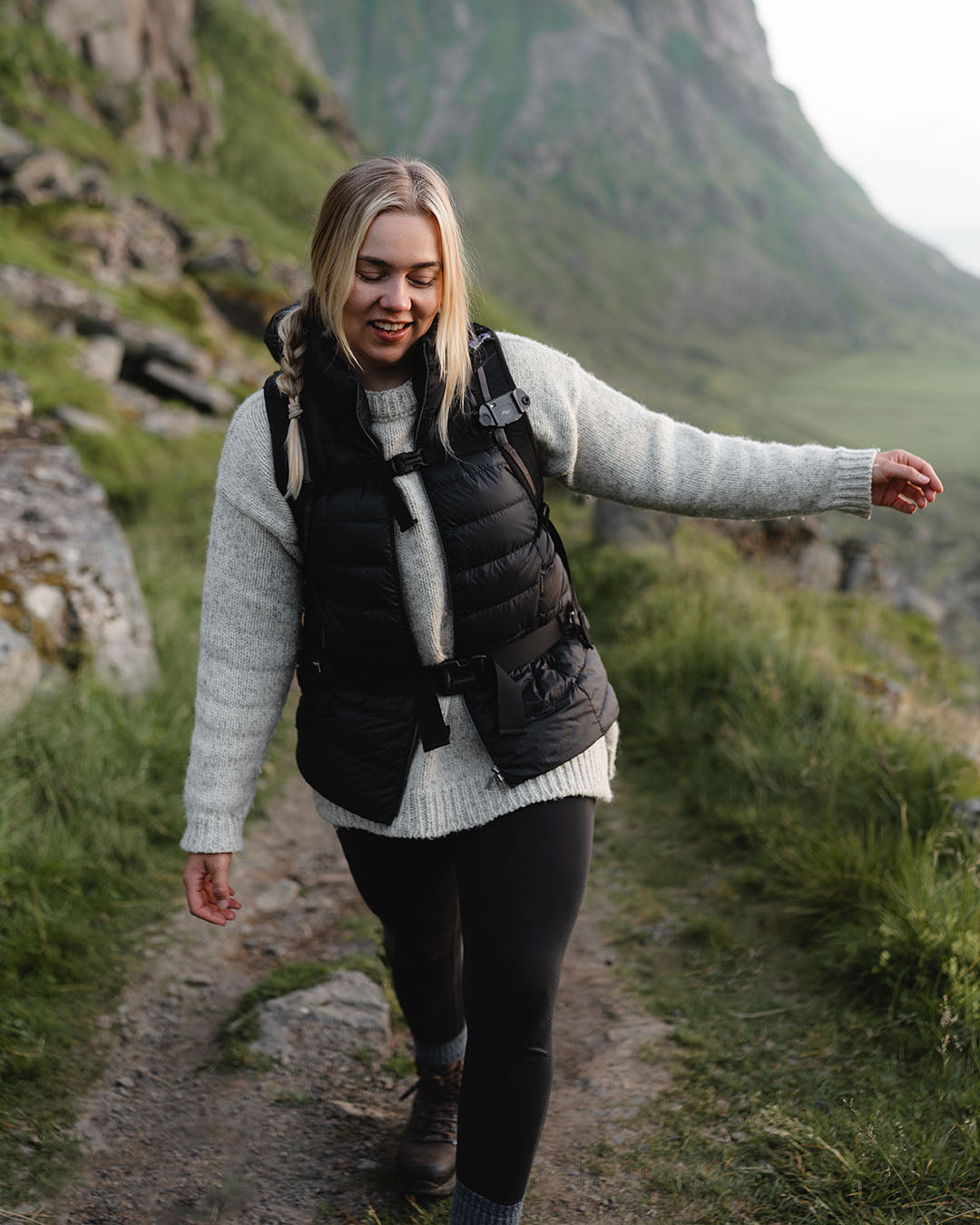 A woman walks in a green mountain landscape, smiling. She is wearing a black Lady down vest (color: charcoal black), a grey knit sweater, and slim black trousers. She carries a backpack on her back.