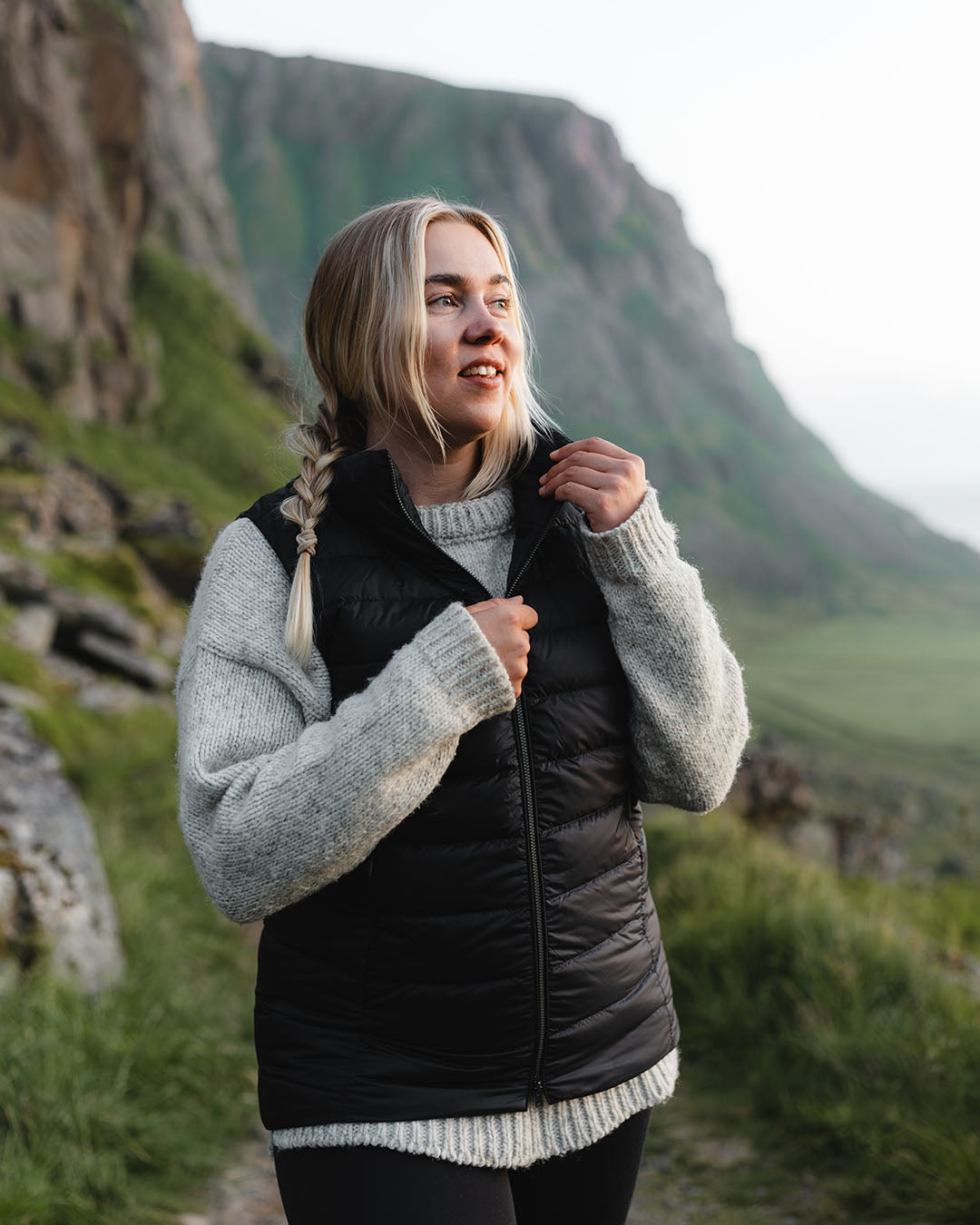 A woman stands in a mountain landscape wearing a black Lady down vest (color: charcoal black) and a grey wool sweater. She looks to the side and holds the zipper of the vest.