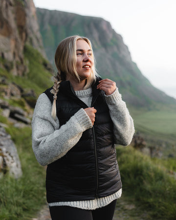 A woman stands in a mountain landscape wearing a black Lady down vest (color: charcoal black) and a grey wool sweater. She looks to the side and holds the zipper of the vest.