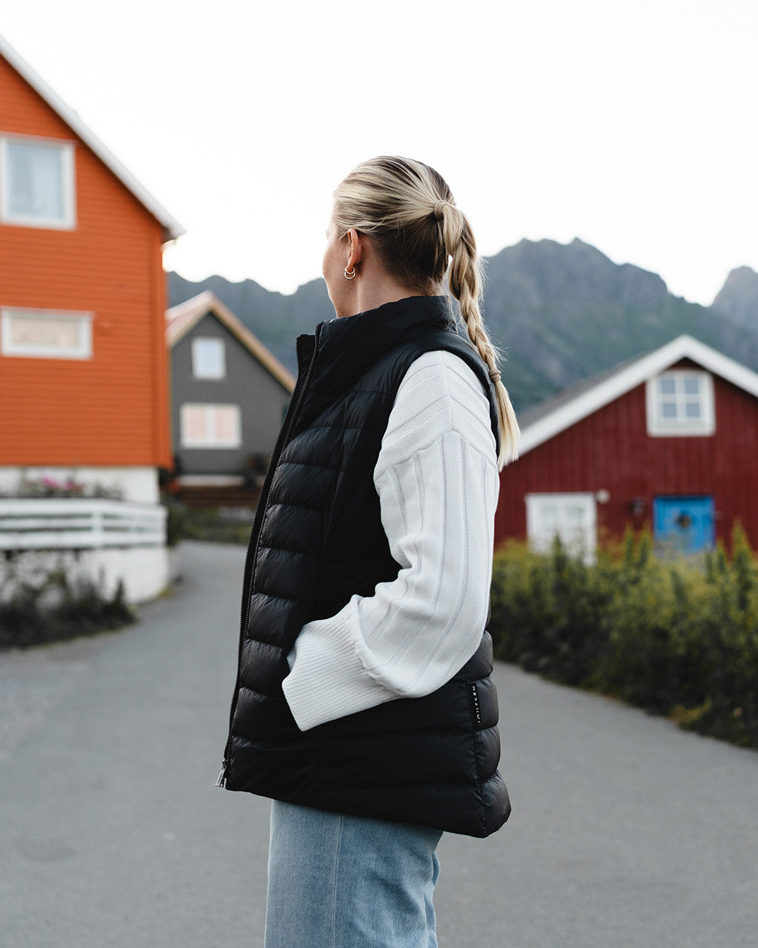 A woman stands in a fishing village landscape, facing the camera sideways. She is wearing a black Lady down vest (color: charcoal black), a white knit sweater, and light blue jeans.