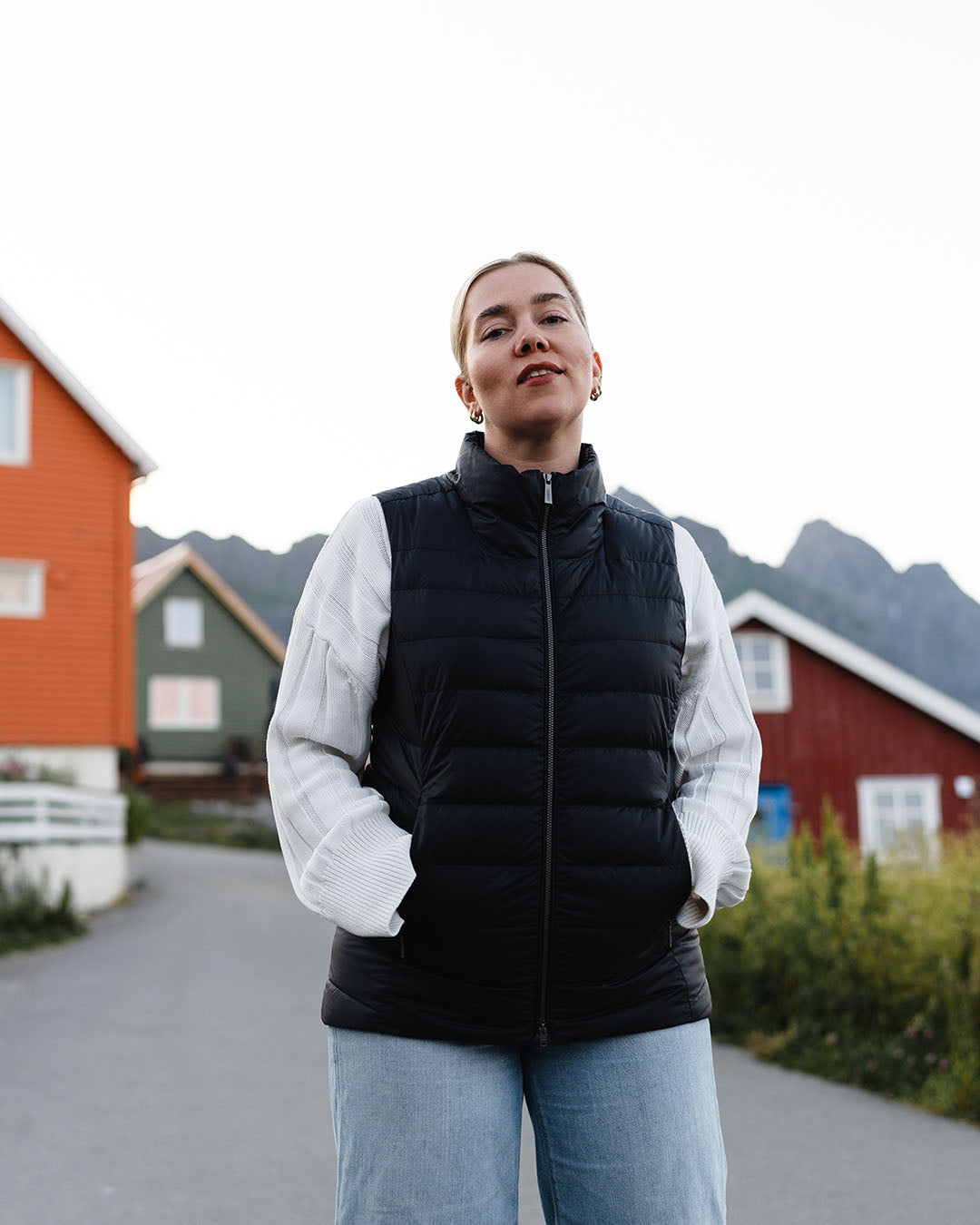 A woman stands in a fishing village, looking at the camera. She is wearing a horizontally quilted black Lady down vest (color: charcoal black), a white knit sweater, and light blue jeans.