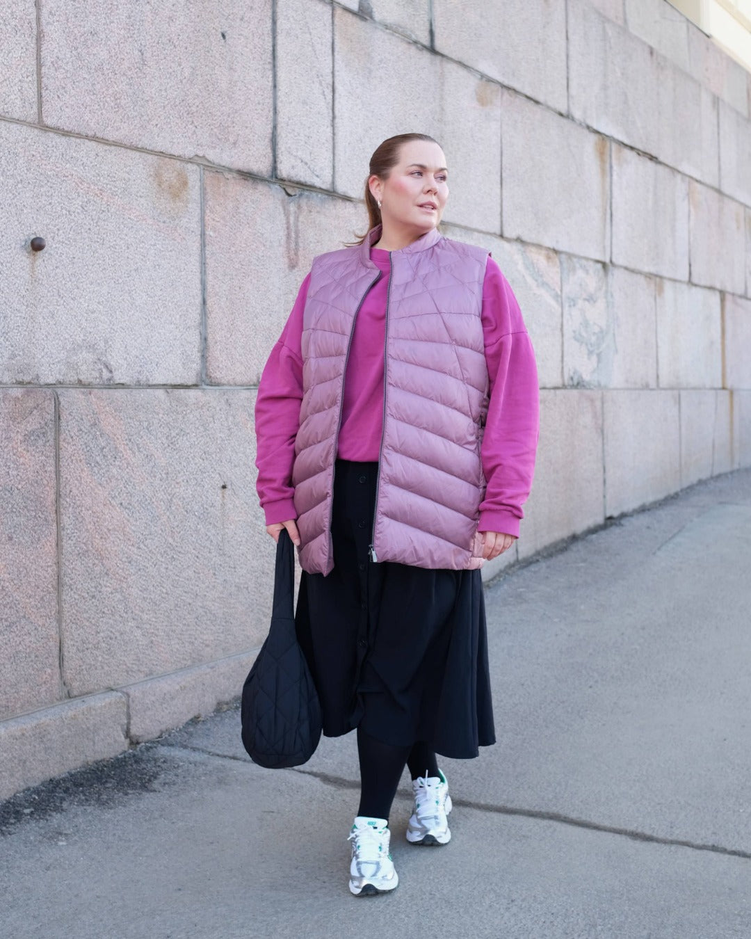 Person wearing a pink down vest over a pink sweater with black pants, standing against a stone wall.