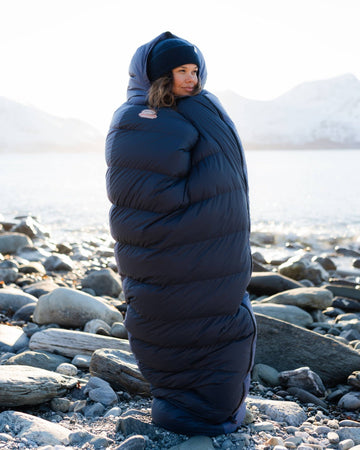 Person wrapped in a large blue sleeping bag on a rocky beach with snow in the background