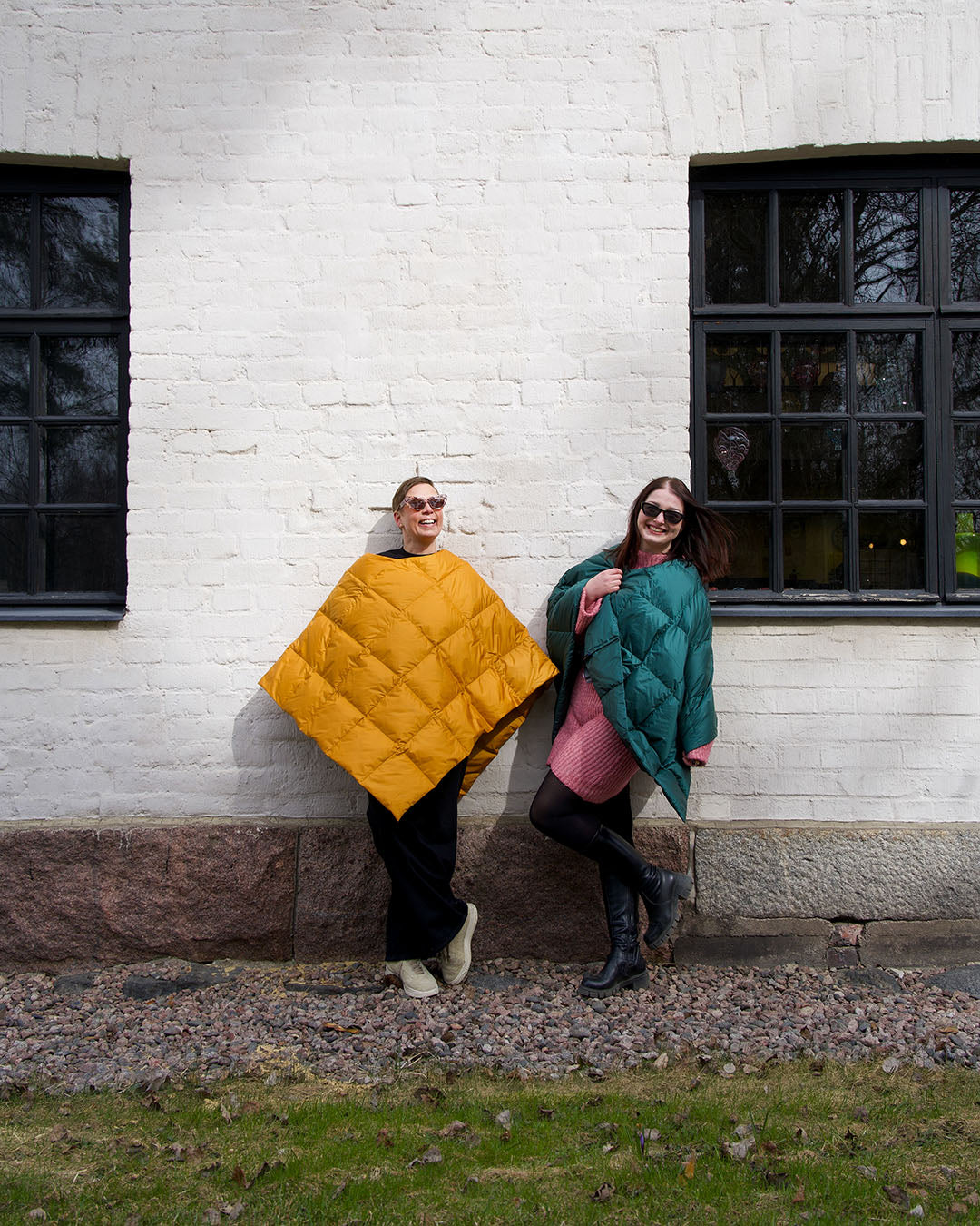 Two people wearing colorful Otava down ponchos standing against a white brick wall.