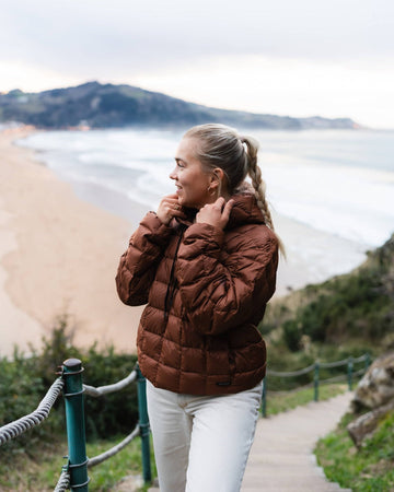 Woman in a brown down hoodie standing on a coastal path with a scenic view.