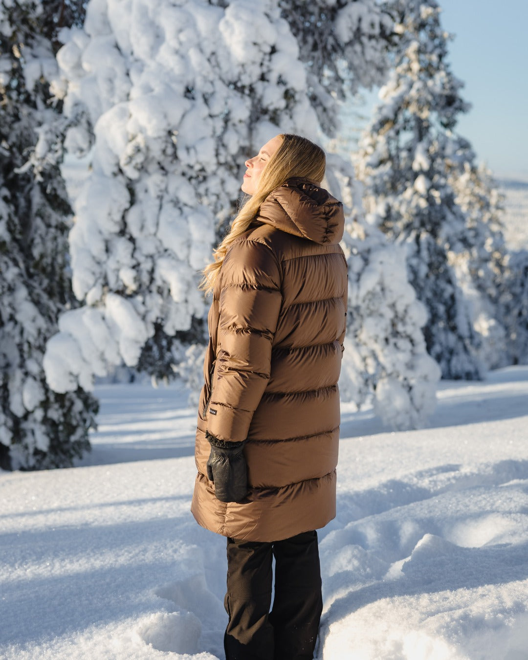 A woman wearing a long brown down coat and black pants stands in a snowy outdoor setting with snow-covered trees in the background.