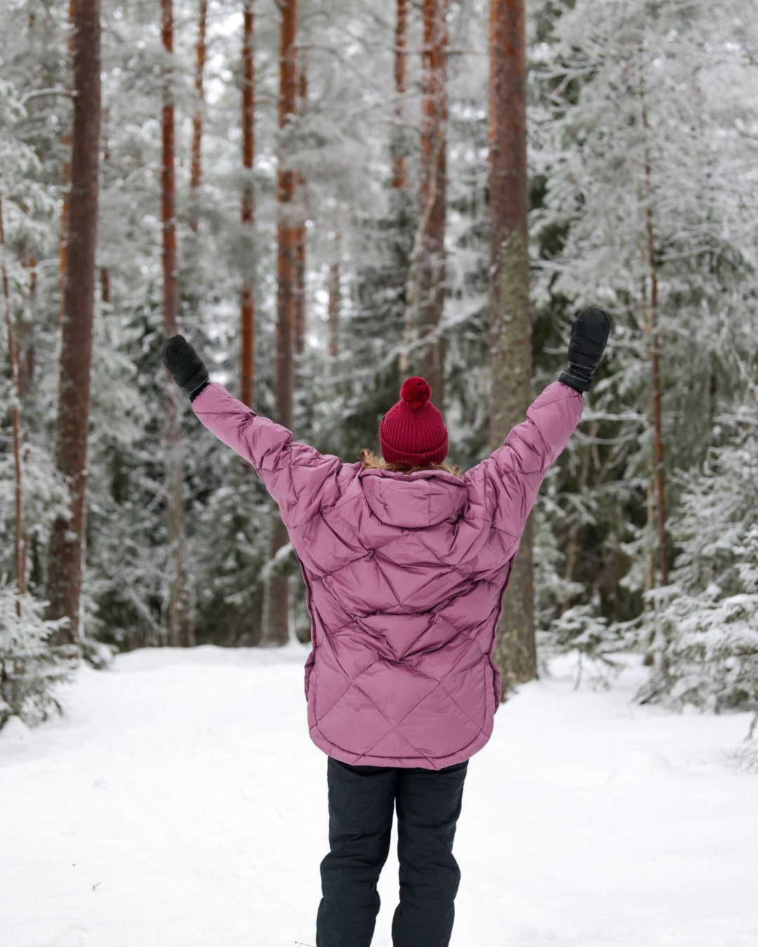 Person in a light pink down anorak and red beanie standing with arms raised in a snowy forest