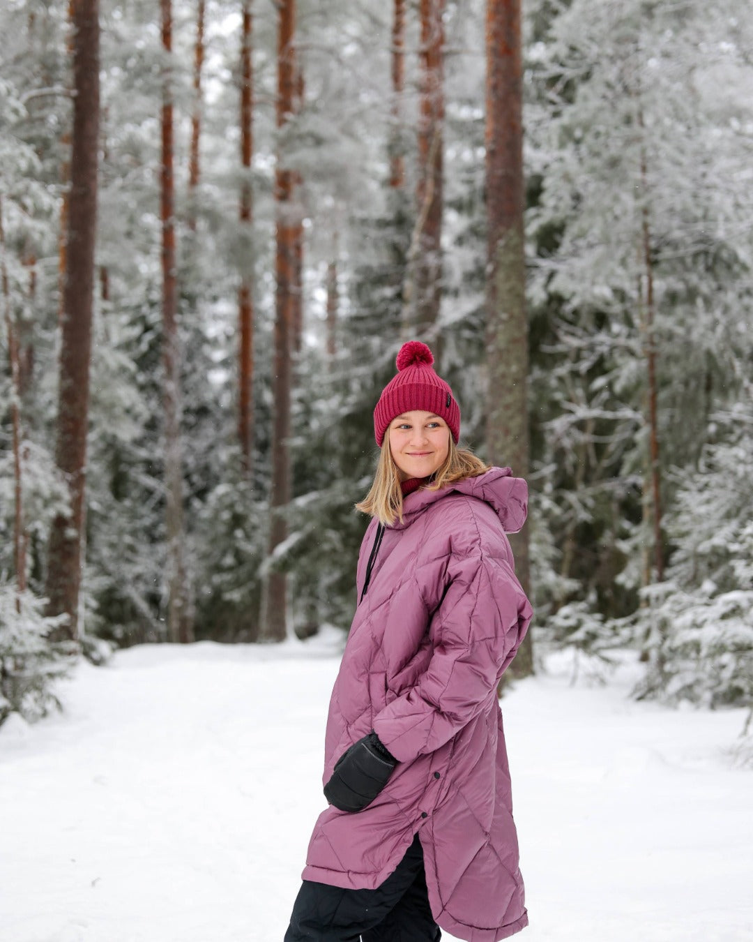 Person wearing a light pink down anorak and hat standing in a snowy forest
