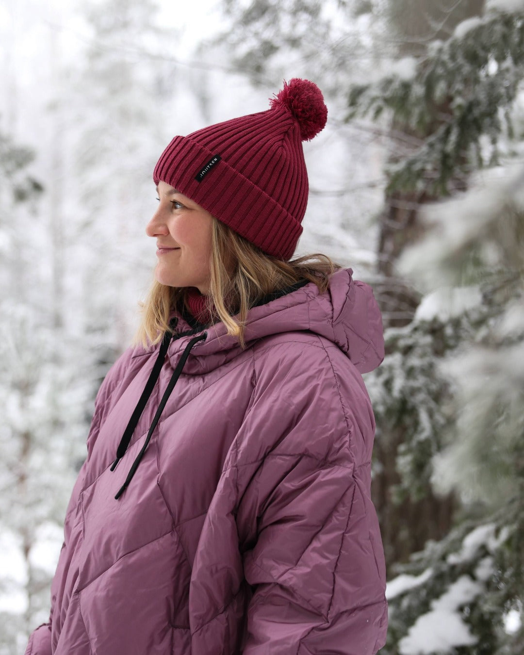 Person wearing a light pink down anorak and red beanie in a snowy forest