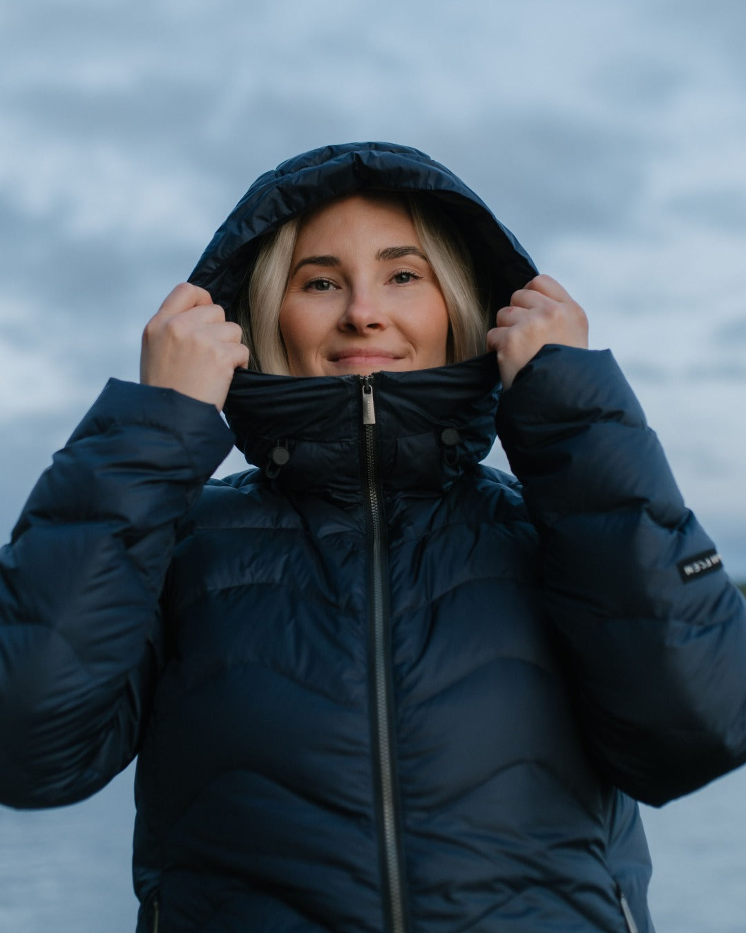 A woman wearing a dark navy blue (color: Night sky) hooded light down coat, holding the hood up around her face against a cloudy, overcast sky.
