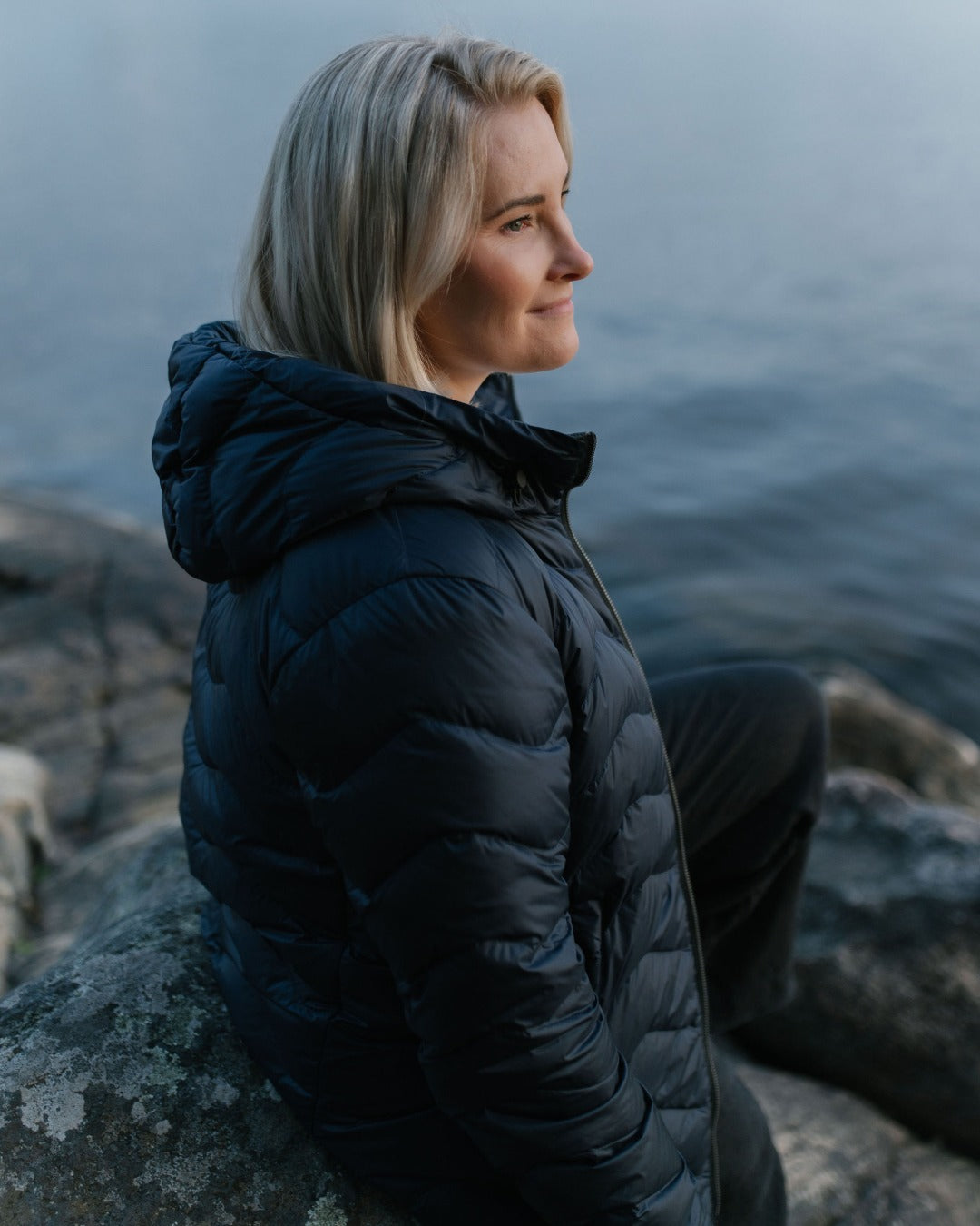 A woman sits on a rock by the water, wearing a navy blue (color: Night sky) light down coat and dark pants, shown in profile against a misty outdoor background.