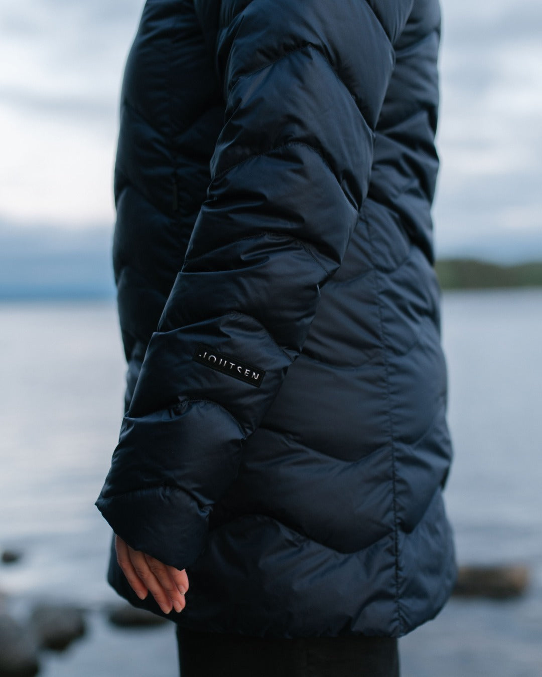 A close-up side view of a person wearing a dark navy (color: Night sky) light down coat outdoors near water, with the sleeve showing a small visible JOUTSEN-brand patch.