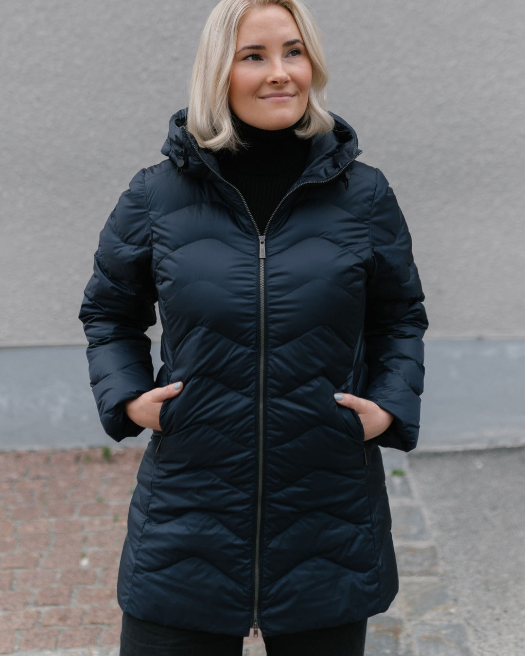 A woman wearing a dark navy blue (color: Night sky) hooded light down coat with a front zipper and pockets, standing outdoors against a gray wall.