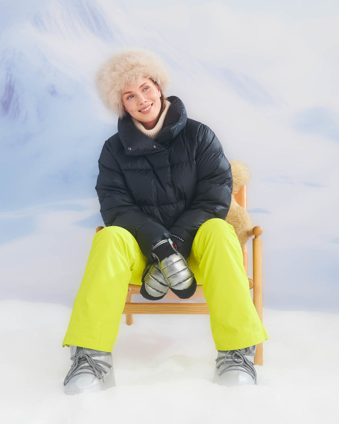 Person sitting on a chair wearing a black puffer jacket and bright yellow pants with a snowy background.