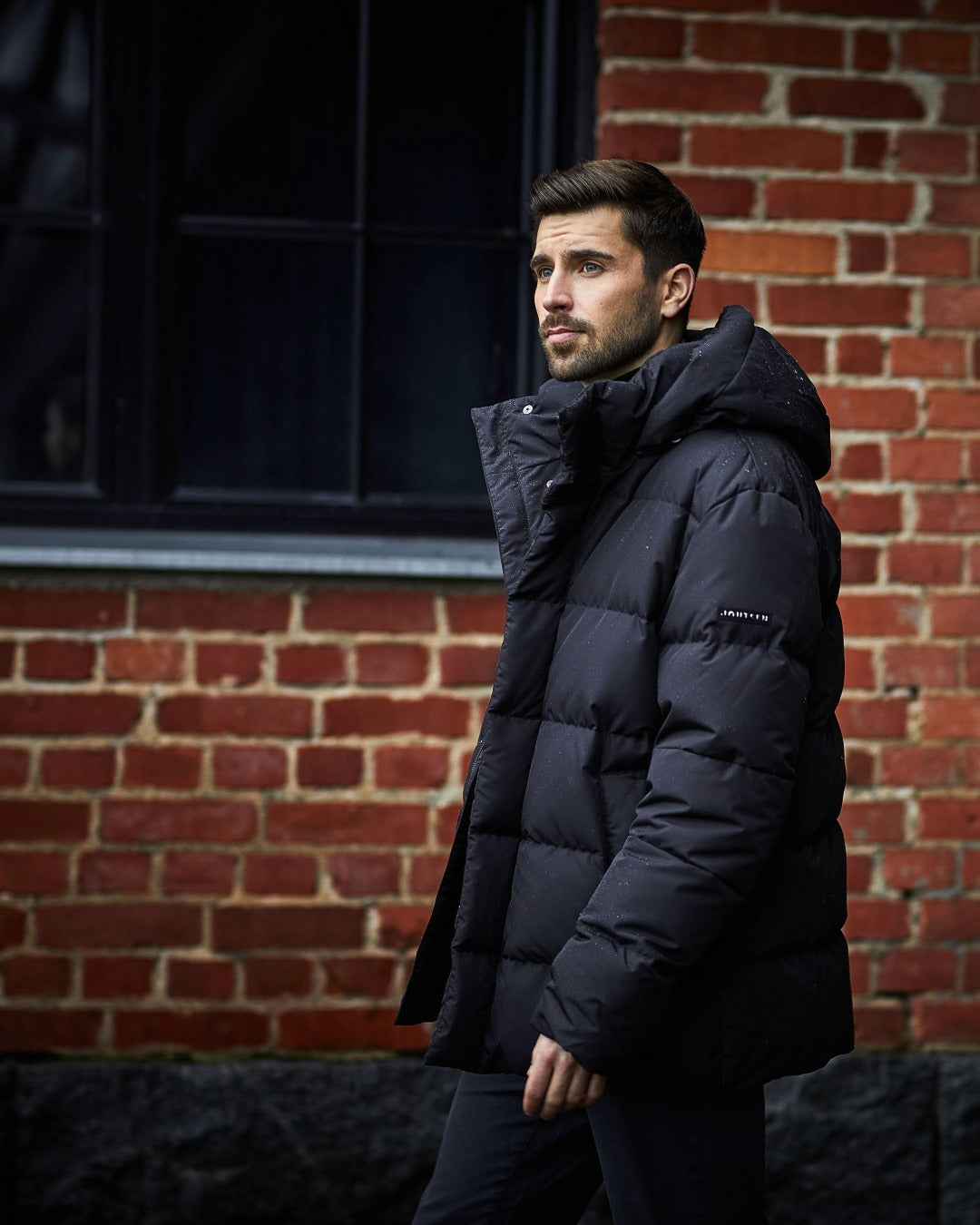 A man wearing a black hooded down coat stands in profile against a red brick wall background.