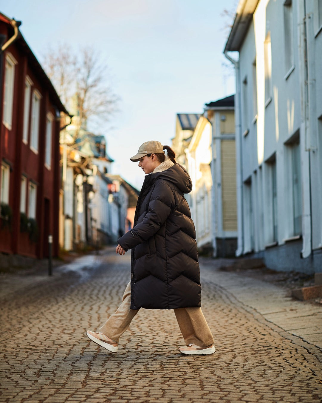 A woman walking down a cobblestone street wearing a long black down coat, beige pants, and a beige baseball cap.