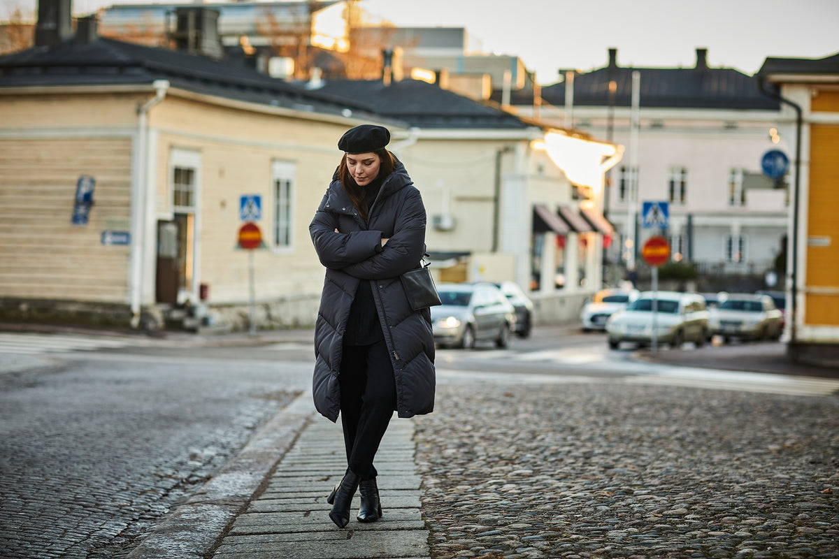 A woman wearing a long black puffer coat, black beret, and black boots stands on a cobblestone street with her arms crossed, looking down. The background shows a softly blurred European-style town street with buildings and parked cars.