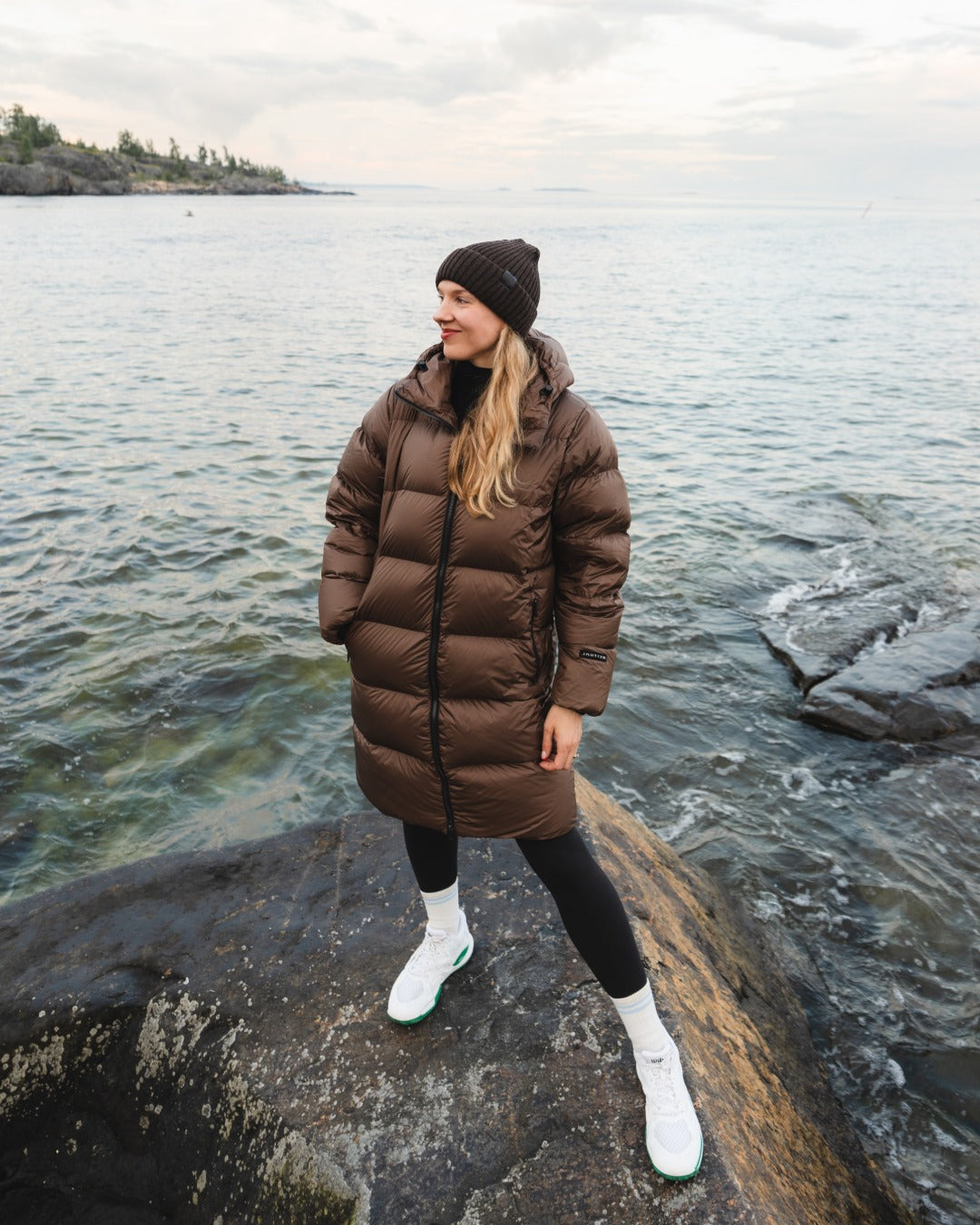 A woman wearing a brown down coat, black beanie, black leggings, and white boots stands on a rocky shoreline with water and an overcast sky in the background.