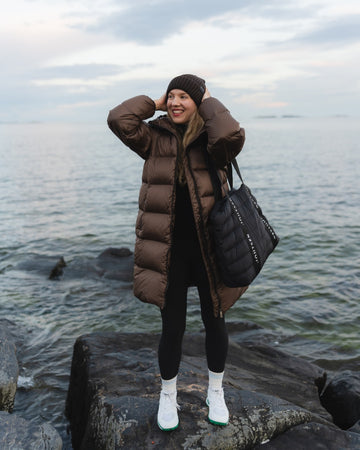 A woman stands on rocks by the ocean wearing a long brown down coat, black beanie, black leggings, white socks, and white sneakers, while carrying a large black tote bag.