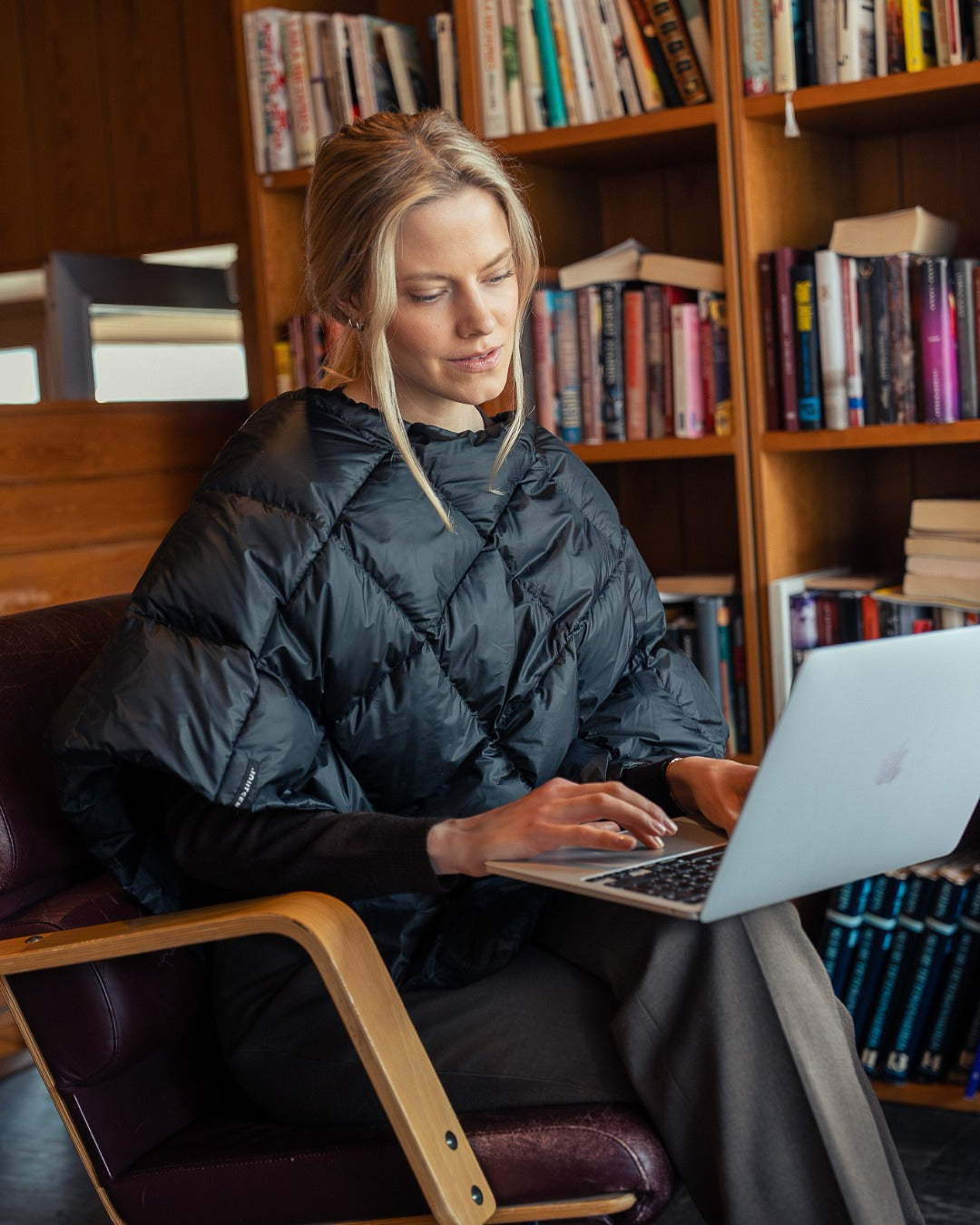 Woman using a laptop in a library setting. She is wearing a black Unelma down poncho.