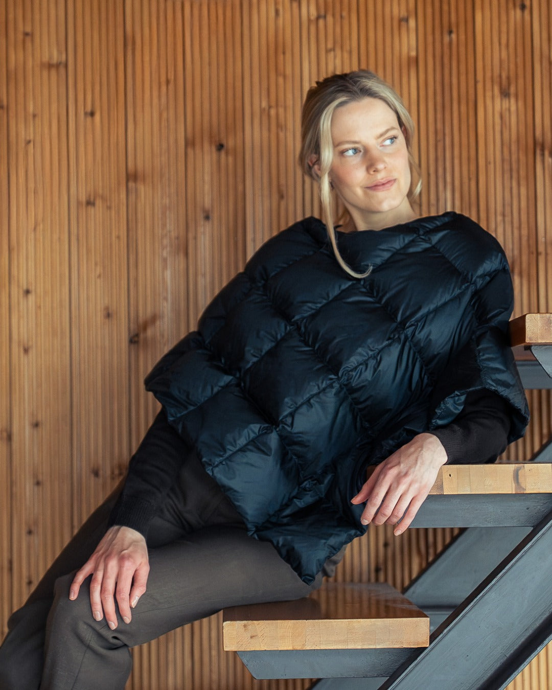 Woman wearing  a black Unelma down poncho sitting on a wooden staircase.