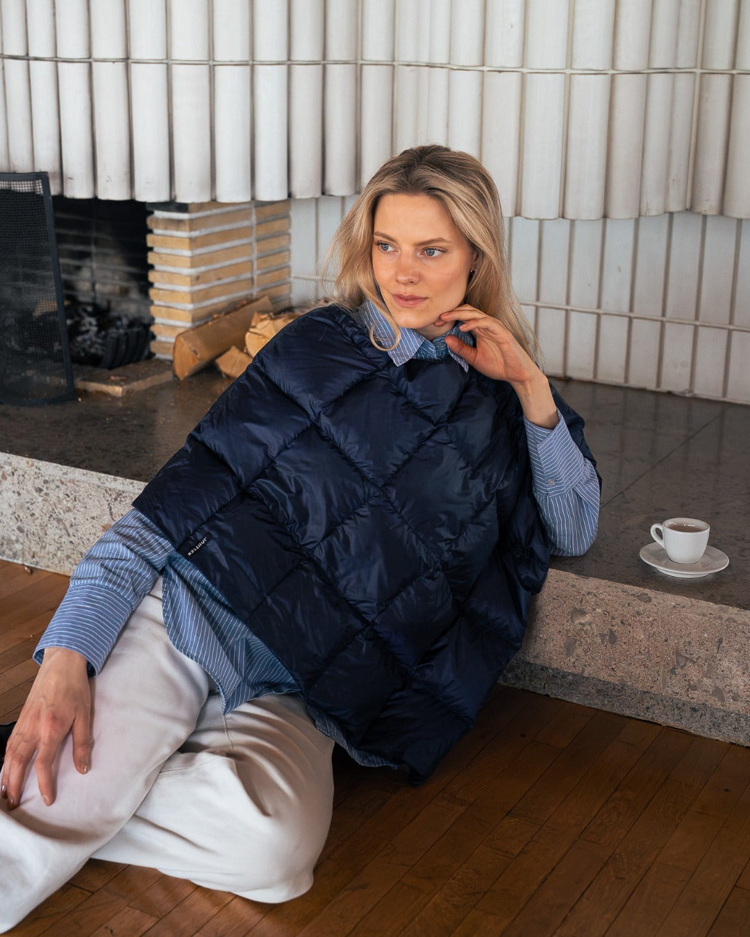 Woman wearing a dark blue Unelma down ponchot sitting by a fireplace.