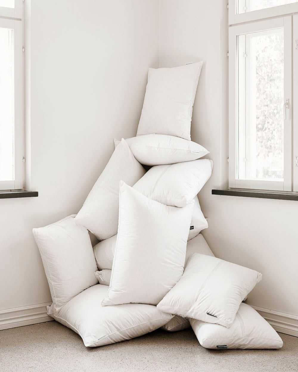 Stack of white down pillows against a white wall with windows.