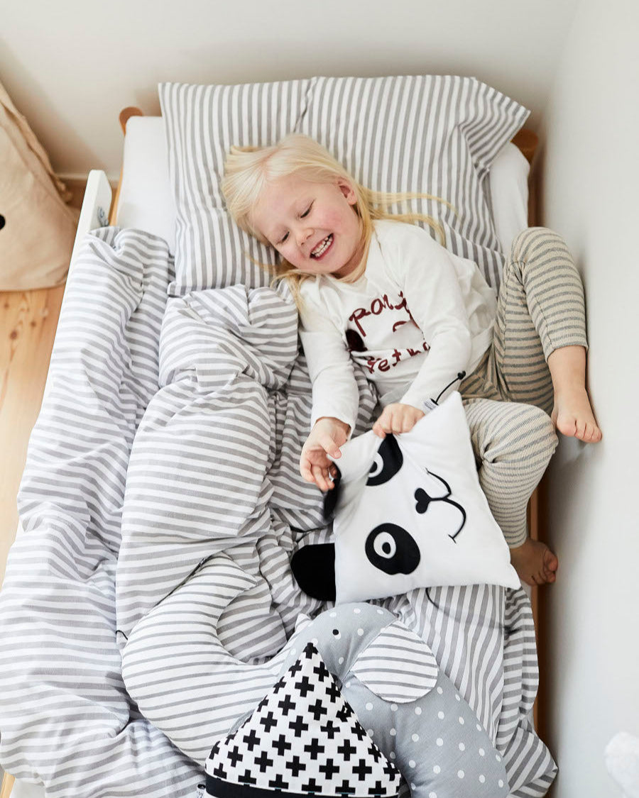 A fair-haired child is lying on a bed with grey and white striped bedding, holding a small panda-print pillow. The bed also has other patterned cushions in black, white, and grey tones.