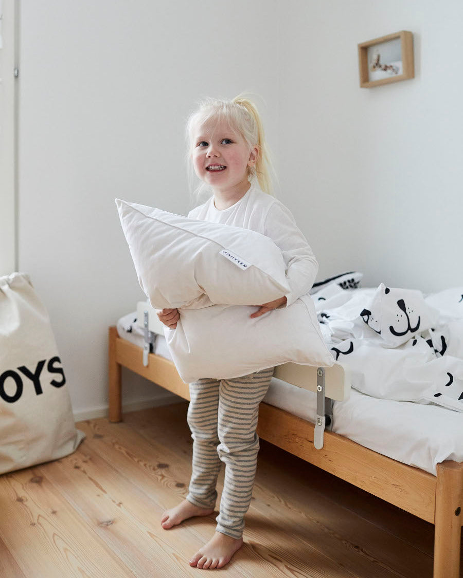 A fair-haired child stands by a wooden children’s bed holding a white pillow. The bed has white sheets with black animal print patterns.