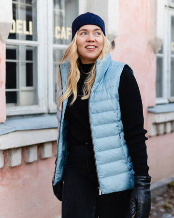 A woman stands on a street wearing a light blue Lady down vest (color: BlueBell). The vest has horizontal quilting and a slight sheen. She is also wearing a black beanie, a turtleneck sweater, trousers, and leather gloves.