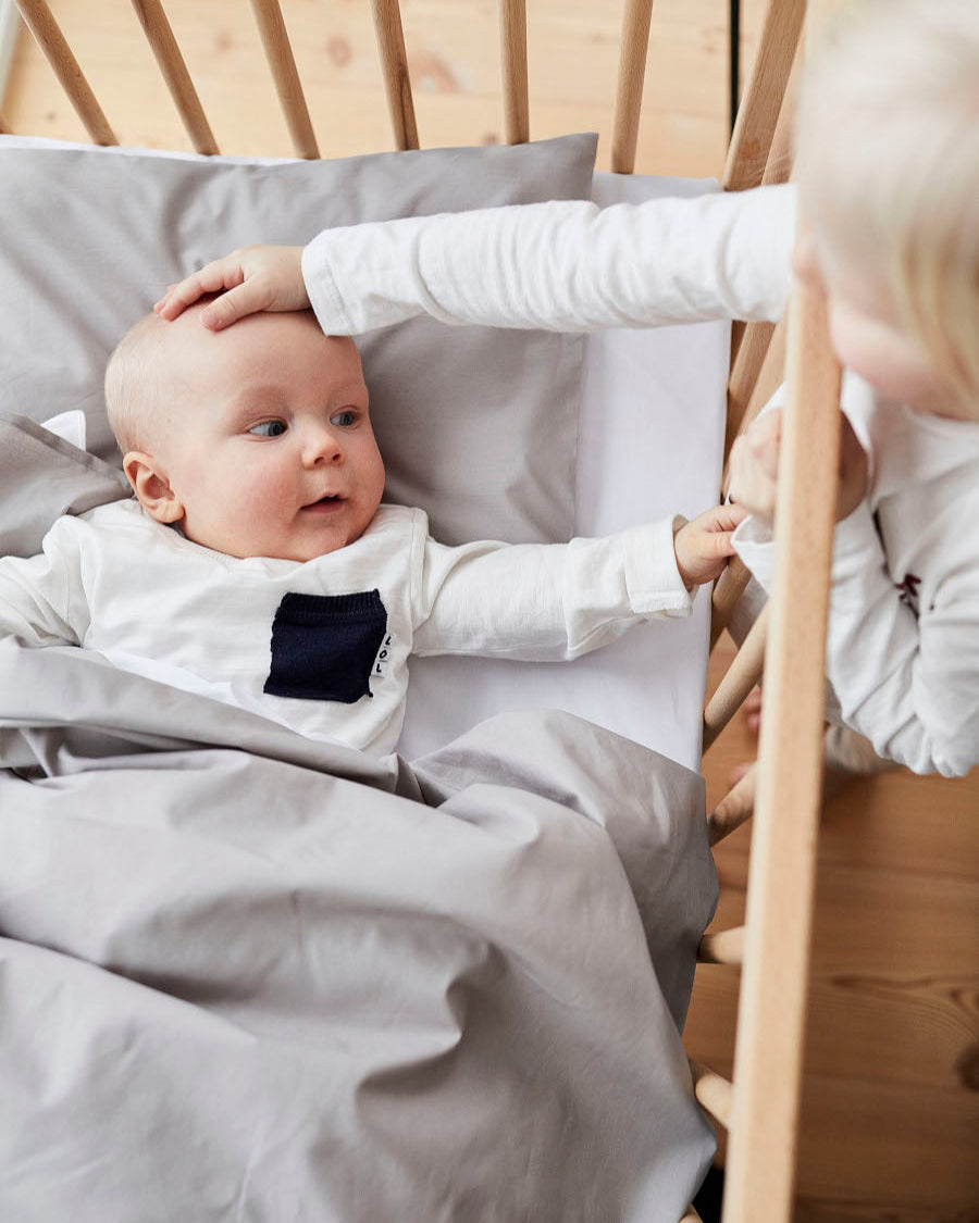 A baby lies in a wooden crib under light grey bedding, looking up while an older child gently touches the baby’s head and hand.