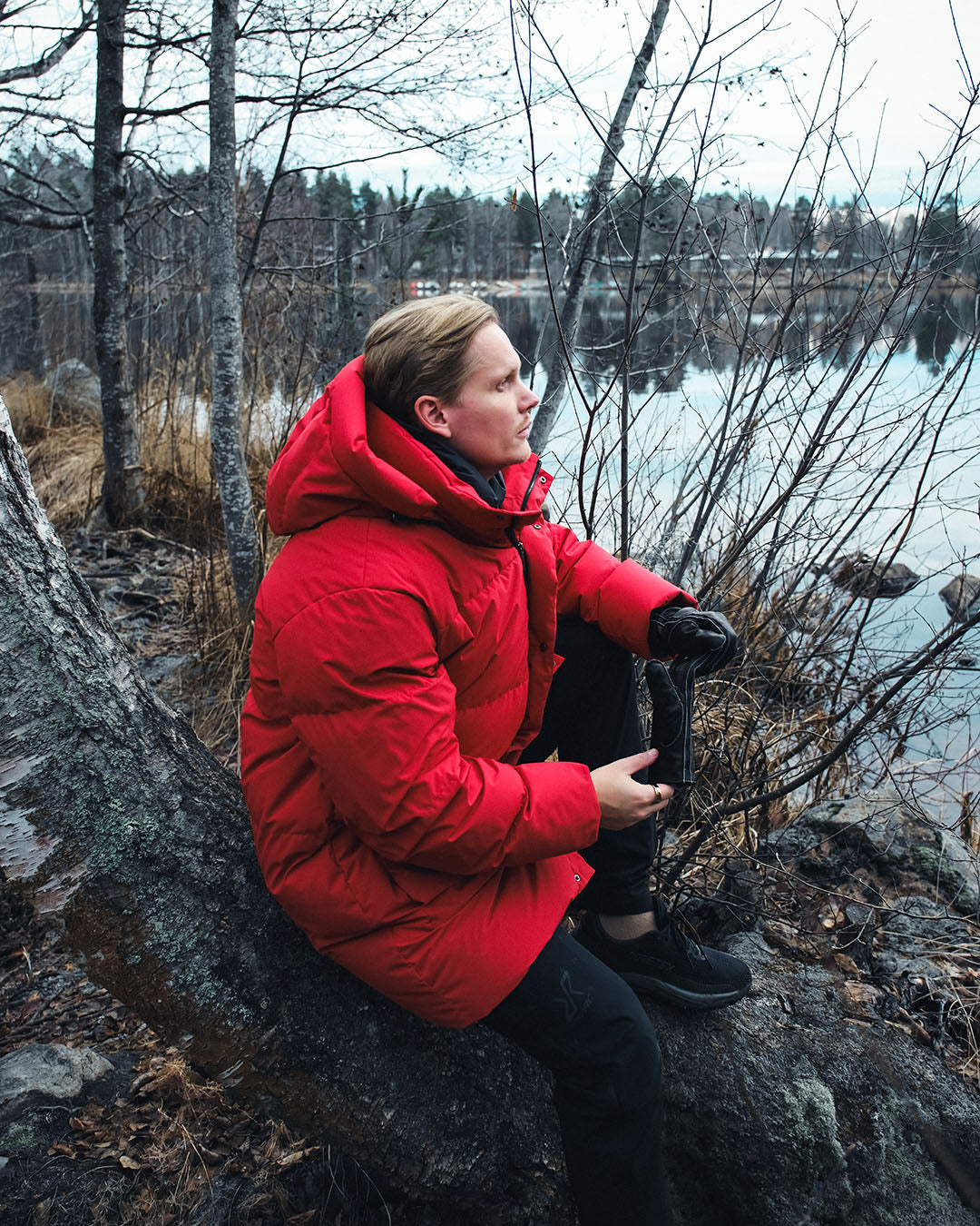 Person in a red down coat sitting on a rock by a lake with bare trees.