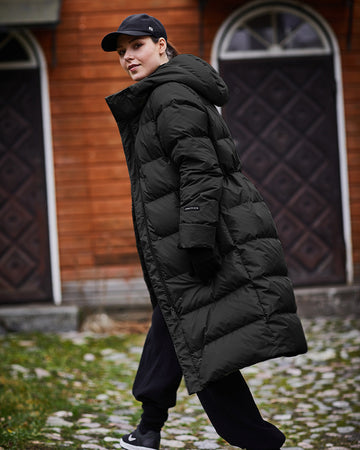 A woman walks outdoors wearing a black, horizontally quilted, long Ulrika down coat, a black cap, and black pants. In the background, there is an old wooden building and a stone-paved yard.