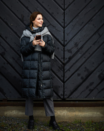 A woman wearing a black, long, slim-fit Essi light down coat (color: charcoal black) with square quilting. Her outfit includes a gray scarf, gray pants, and black shoes. She is holding a phone, and a black wooden door is visible in the background.