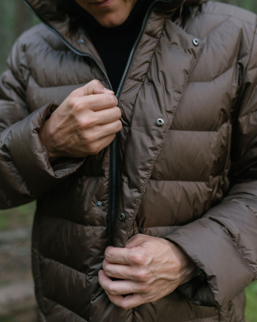 A close-up of a person wearing a brown down coat and zipping it, with a dark turtleneck underneath. The background is a blurred outdoor forest setting.