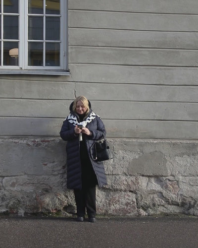 A woman is walking beside a wall, smiling. She is wearing a dark blue Säde light down coat (color: Night sky), black clothes and accessories. A striped knit is tied around her shoulders.