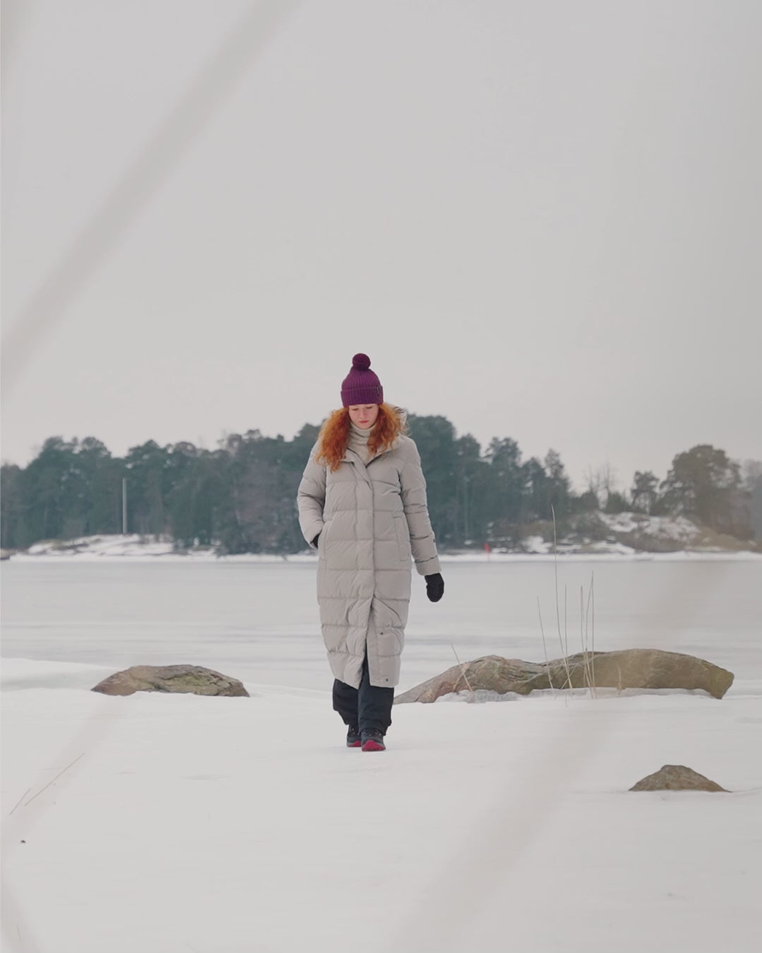 A woman is walking in a winter environment on the shore, wearing Joutsen’s long light-colored hooded Blanka down coat (color: sand dune). She has a purple pom-pom beanie on her head and sneakers on her feet.