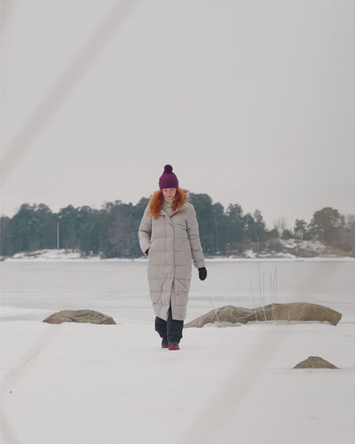 A woman is walking in a winter environment on the shore, wearing Joutsen’s long light-colored hooded Blanka down coat (color: sand dune). She has a purple pom-pom beanie on her head and sneakers on her feet.