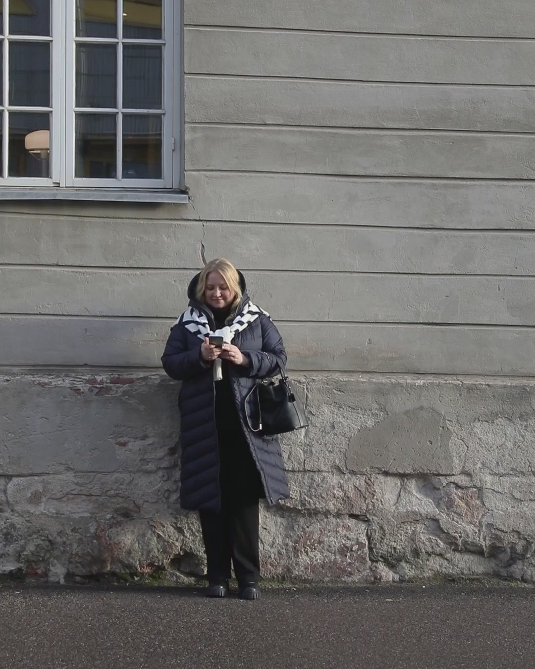 A woman is walking beside a wall, smiling. She is wearing a dark blue Säde light down coat (color: Night sky), black clothes and accessories. A striped knit is tied around her shoulders.