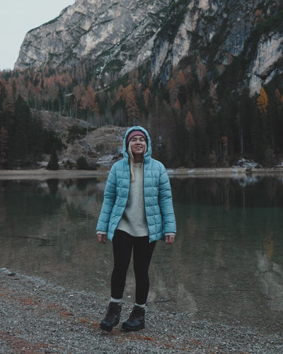 A woman is walking by a lake wearing a light blue light down coat and lilac beanie.