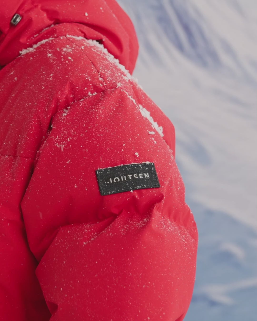 A man is standing in a snowy landscape, putting gloves to his pocket and closes the zipper. He is wearing a long, red (color: chili pepper) Tuisku down parka coat with a large hood. 