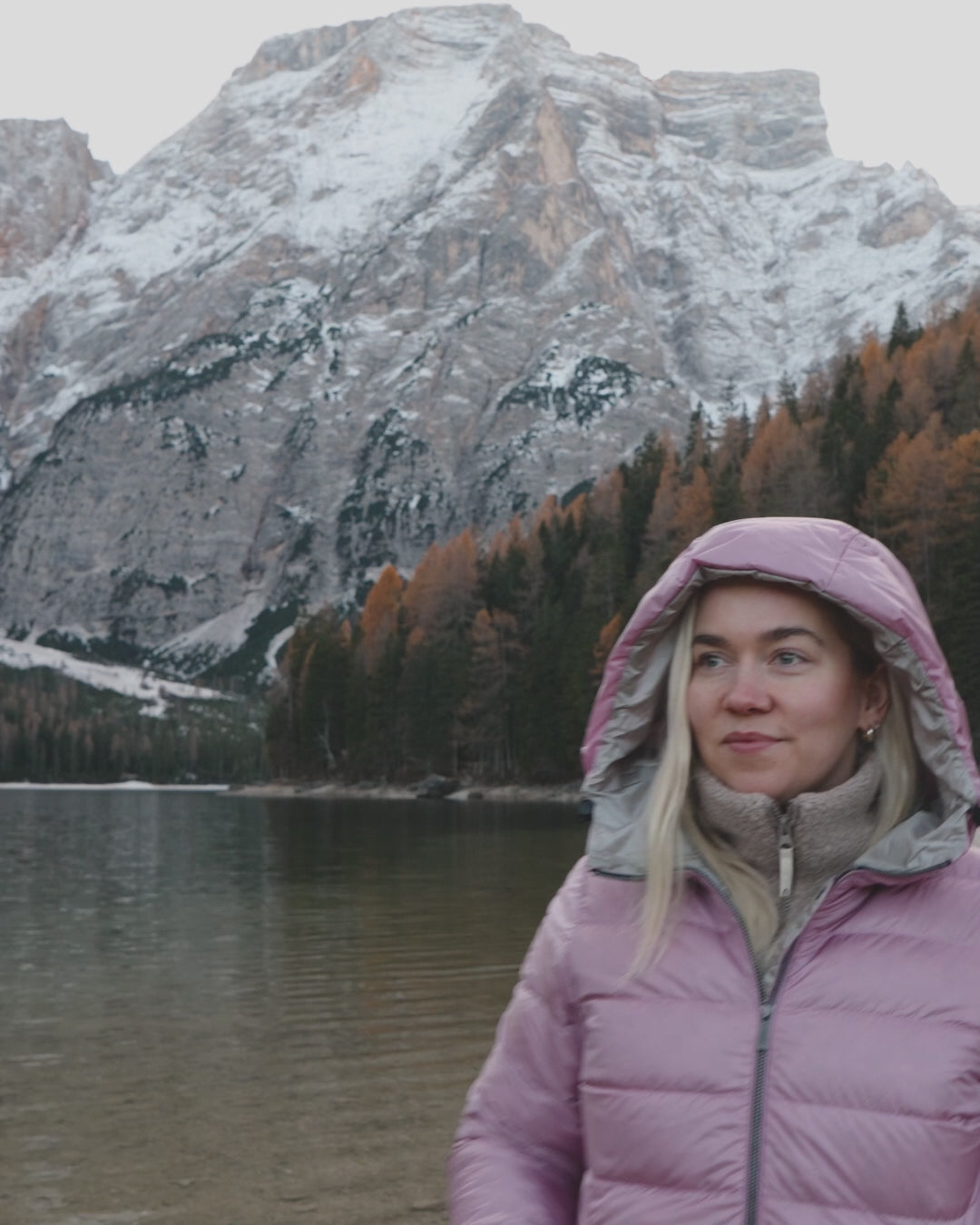 A woman is walking by a lake, wearing light pink light down coat. There are mountains and forest behind her.
