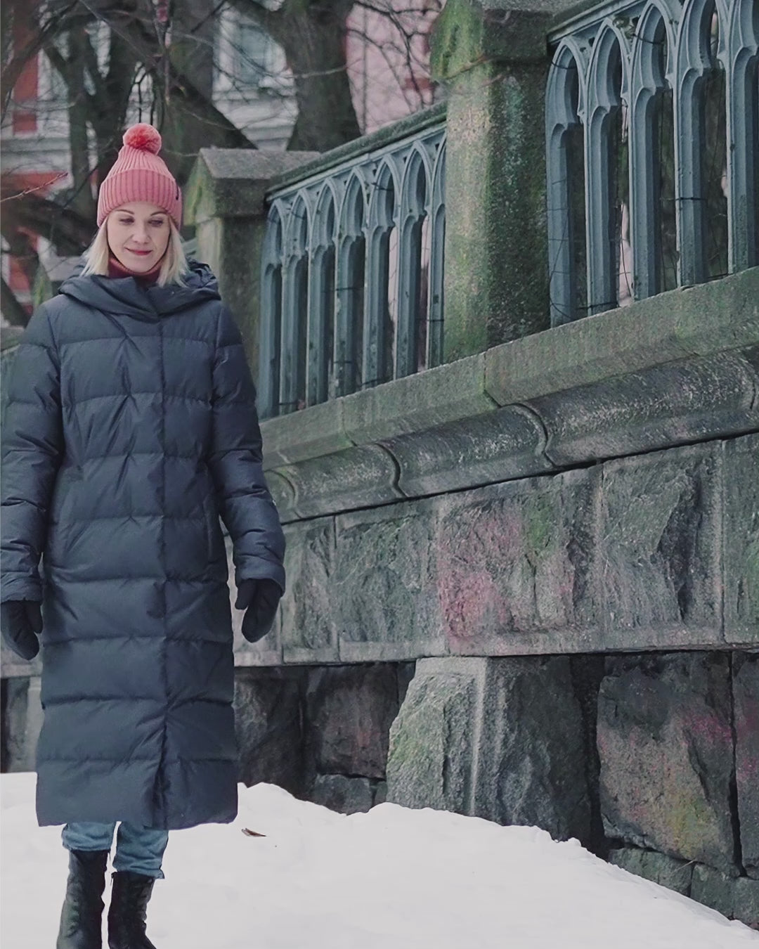 A woman is looking to the side and walking in a winter city environment, wearing Joutsen’s long black Blanka down coat (color: llava). She has a pink beanie on her head and black winter boots on her feet. A stone railing is visible in the background.