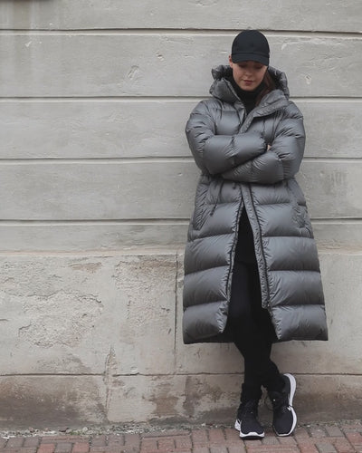 A smiling woman walks in an urban setting wearing a gray (color name: Grey dove) Estrid down coat. The coat’s zipper is open, and it is cinched at the waist with a drawstring.