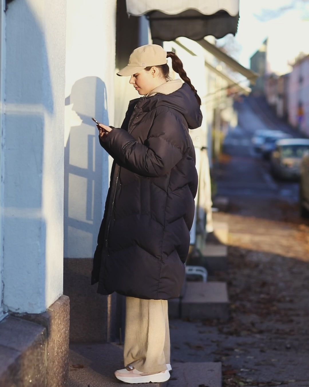 A woman is talking on the phone while walking in the city. She is wearing a long, black Reina down coat. At first, she has paired the coat with light-colored clothes and accessories, and in the end, she is wearing it with black clothes and accessories.