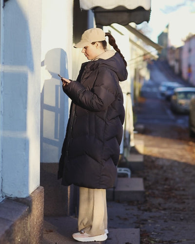 A woman is talking on the phone while walking in the city. She is wearing a long, black Reina down coat. At first, she has paired the coat with light-colored clothes and accessories, and in the end, she is wearing it with black clothes and accessories.