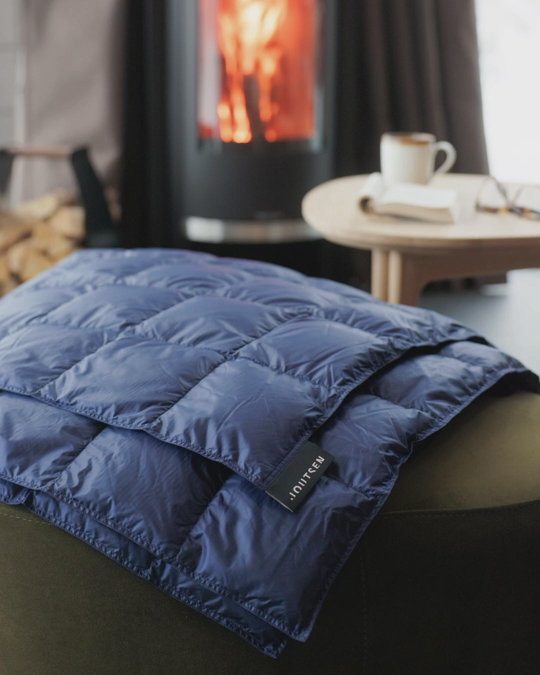 A blue (color: Honeyberry) Kulkuri down balnket is placed on a sofa. There is a fireplace in the background