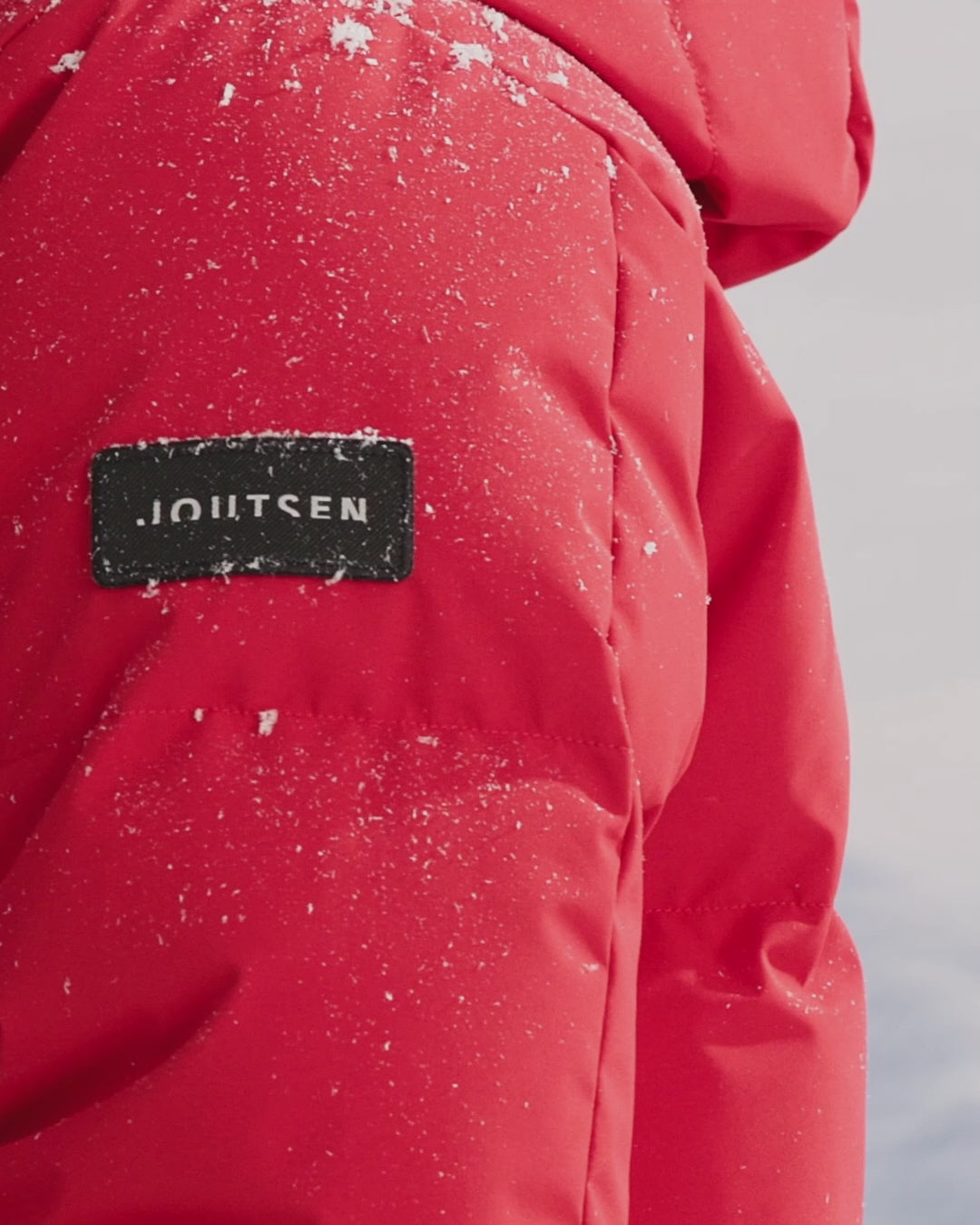 A woman is standing in a snowy landscape, blowing snow from her hands. She is wearing a long, red (color: chili pepper) Siiri down parka coat with a large hood. 
