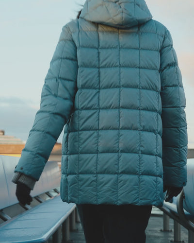 A woman is walking in a boat away from the camera, wearing a quilted light blue Tulppaani light down coat (color: Blue bell) and a brown beanie.