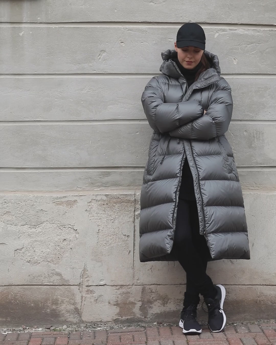 A smiling woman walks in an urban setting wearing a gray (color name: Grey dove) Estrid down coat. The coat’s zipper is open, and it is cinched at the waist with a drawstring.